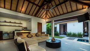 A modern living area with a sofa, coffee table, and kitchen in the background. - Arama Riverside (Legian)