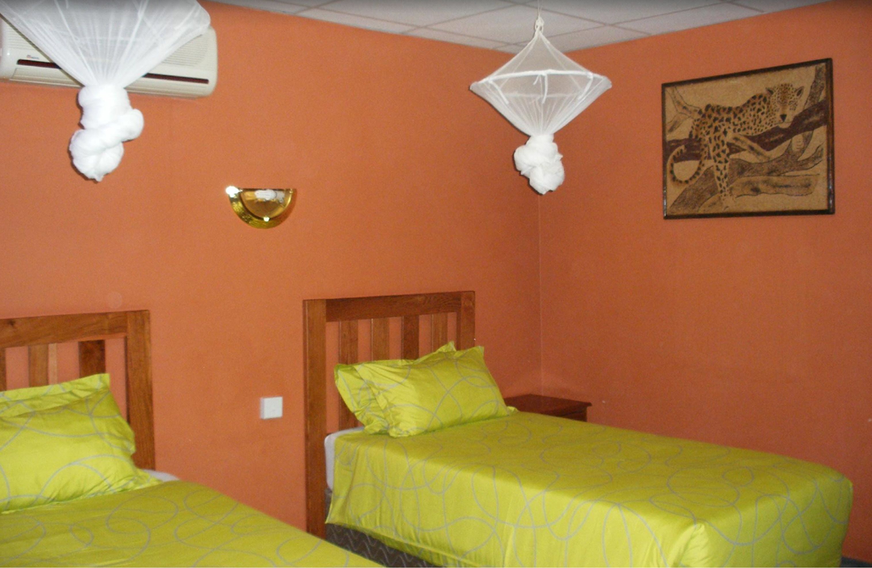A hotel room with two beds, each with a green bedspread and matching pillows. There is a wall-mounted air conditioner and a decorative piece above the beds.
