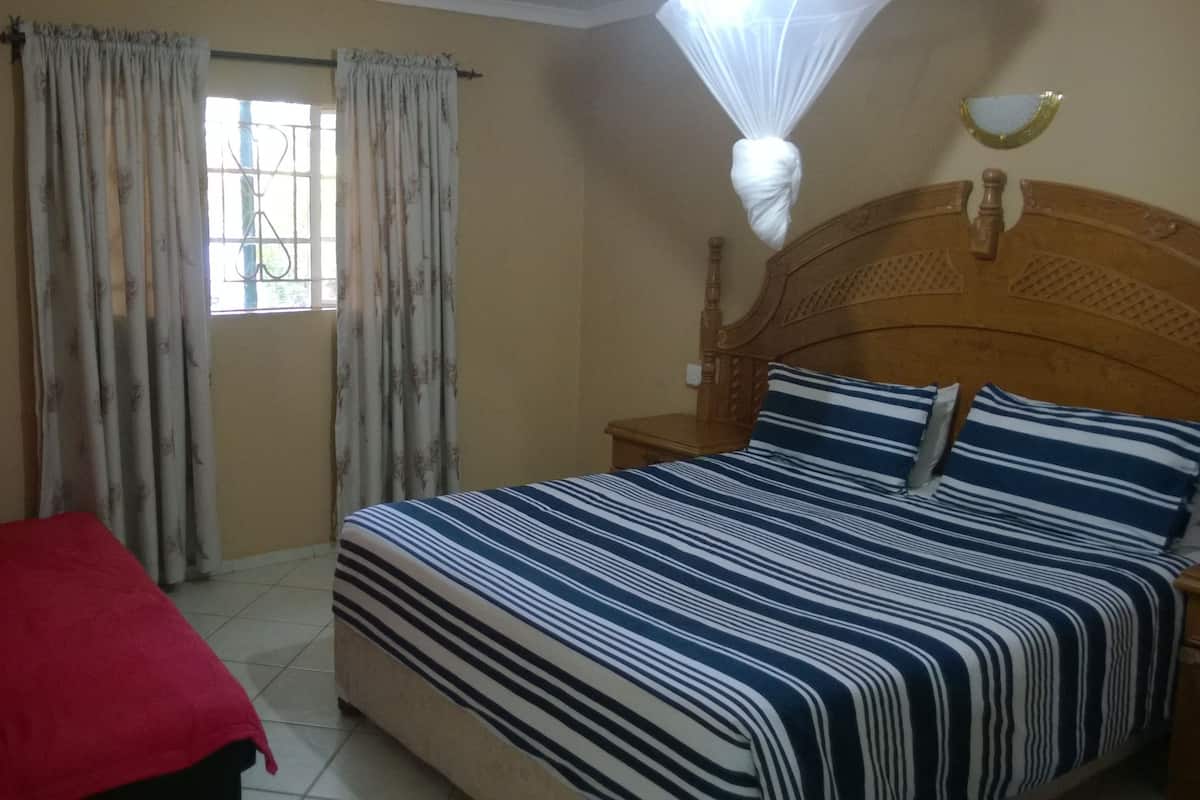 A bedroom with a bed, a nightstand, a chair with a red cover, and a window with curtains.