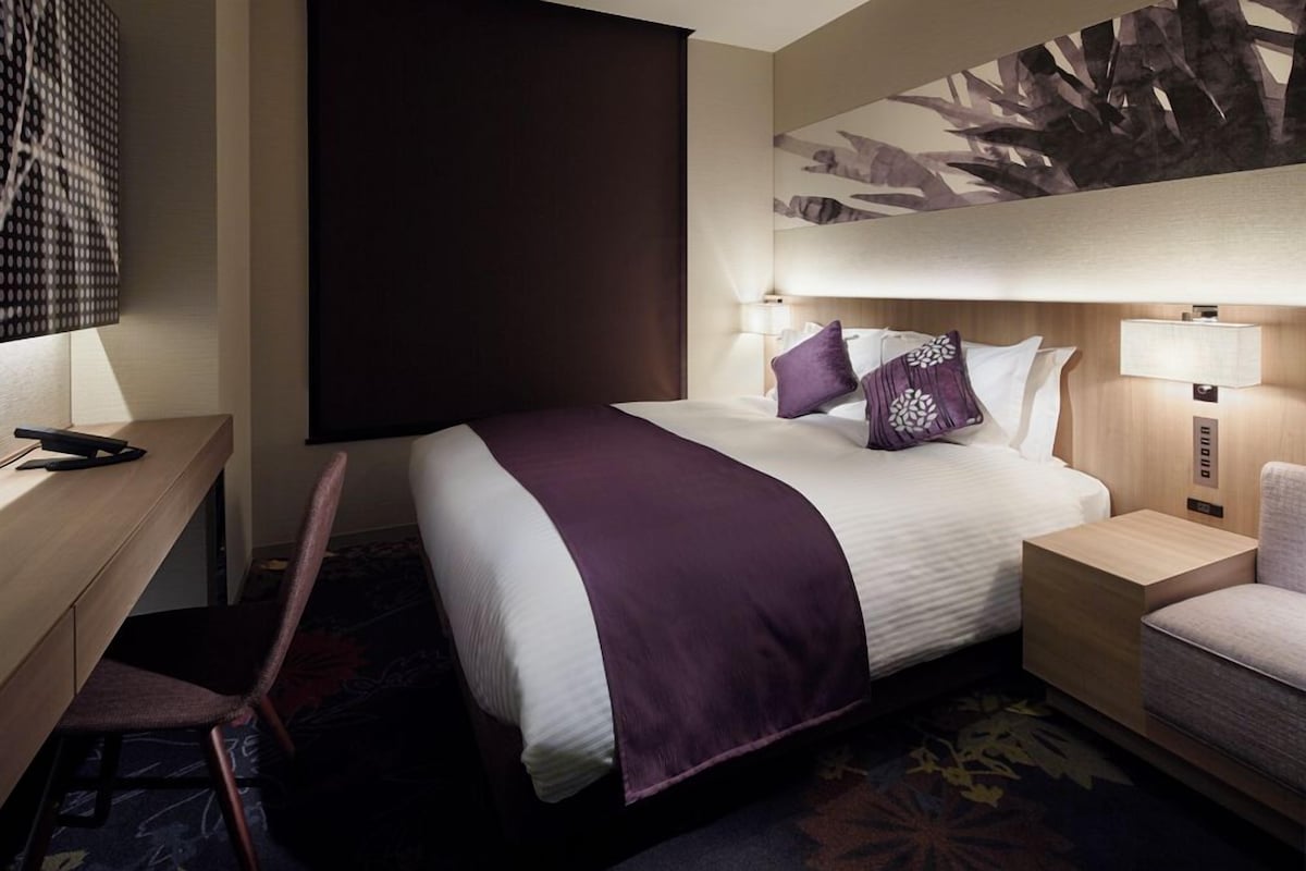 A hotel room with a bed, bedside tables, a desk, a chair, and a wall-mounted artwork.