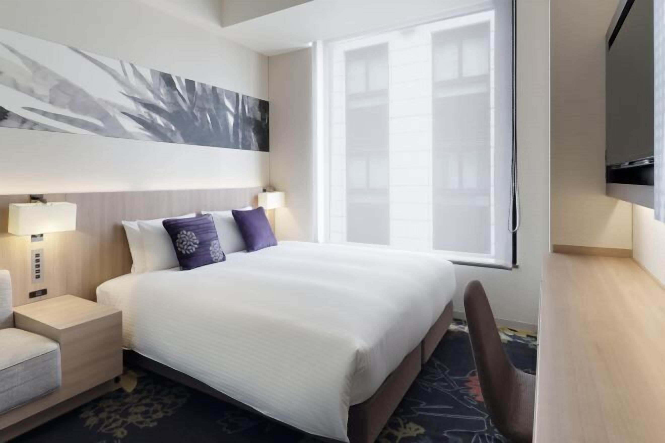 Een hotelkamer met een groot bed, bedlampjes, een flatscreen televisie en een raam met jaloezieën.