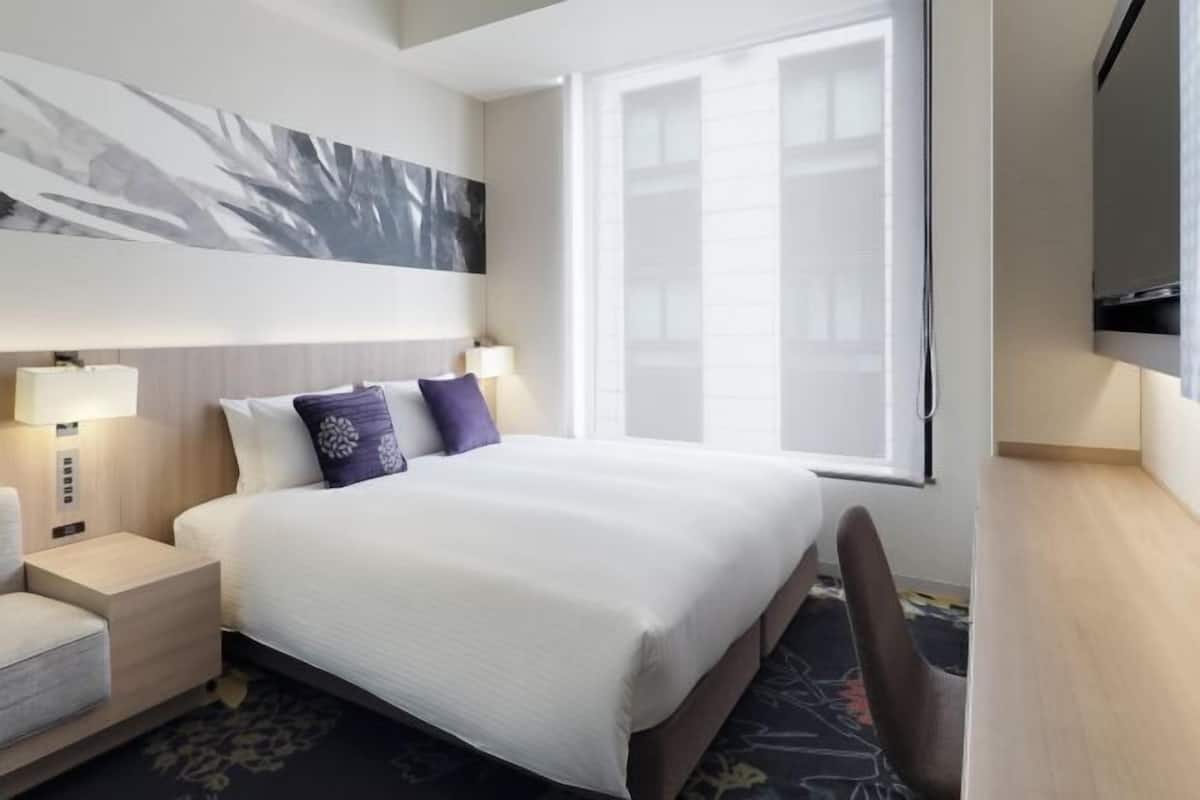 A hotel room with a large bed, bedside lamps, a flat-screen TV, and a window with blinds.