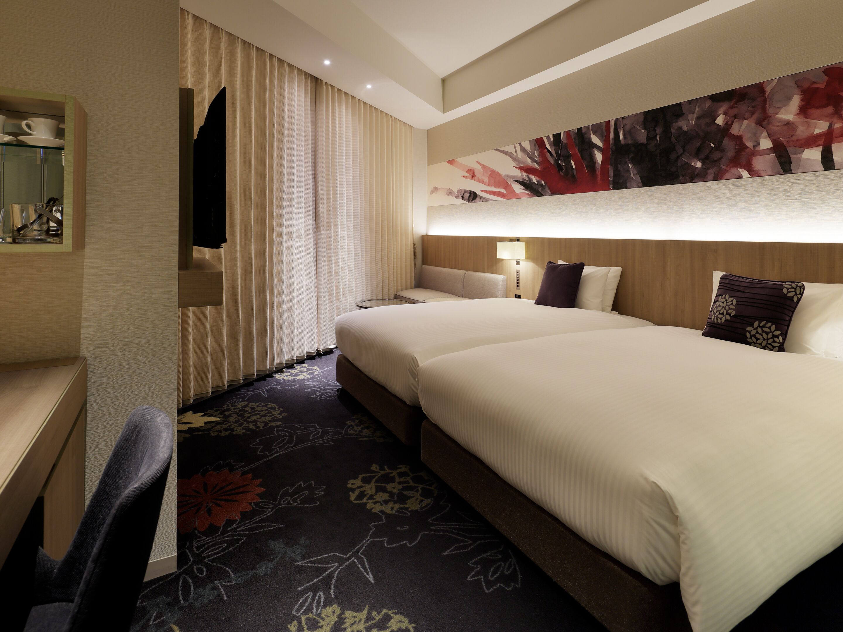 A hotel room with a large bed, a desk, a chair, a TV, and a wall-mounted lamp.