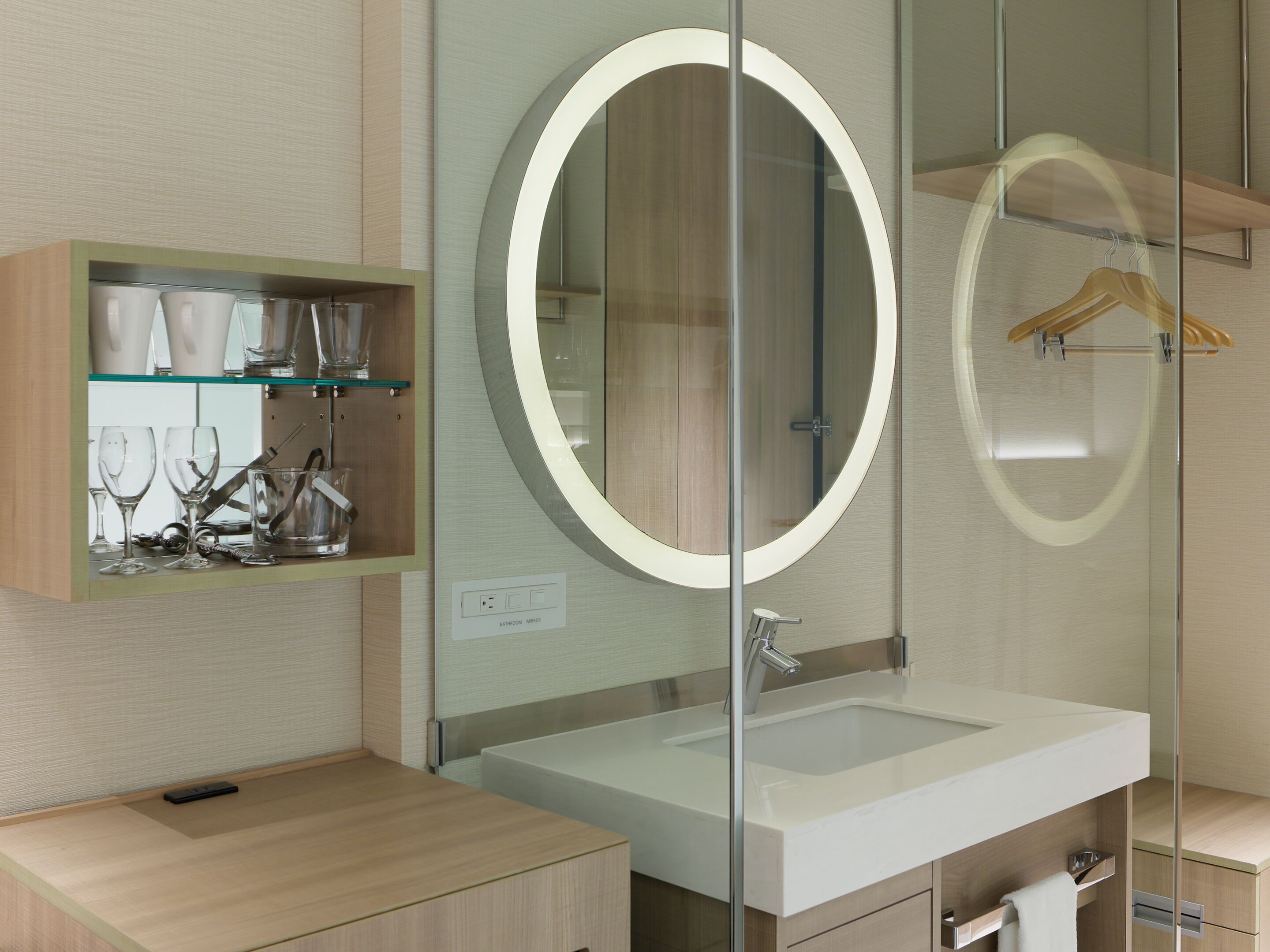 Un baño moderno con tocador de vidrio, un espejo redondo con luz y un estante de madera con vasos.