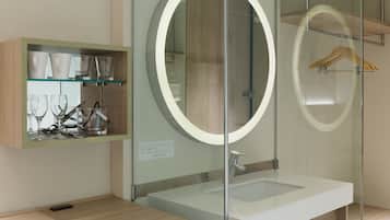 Un baño moderno con tocador de vidrio, un espejo redondo con luz y un estante de madera con vasos.