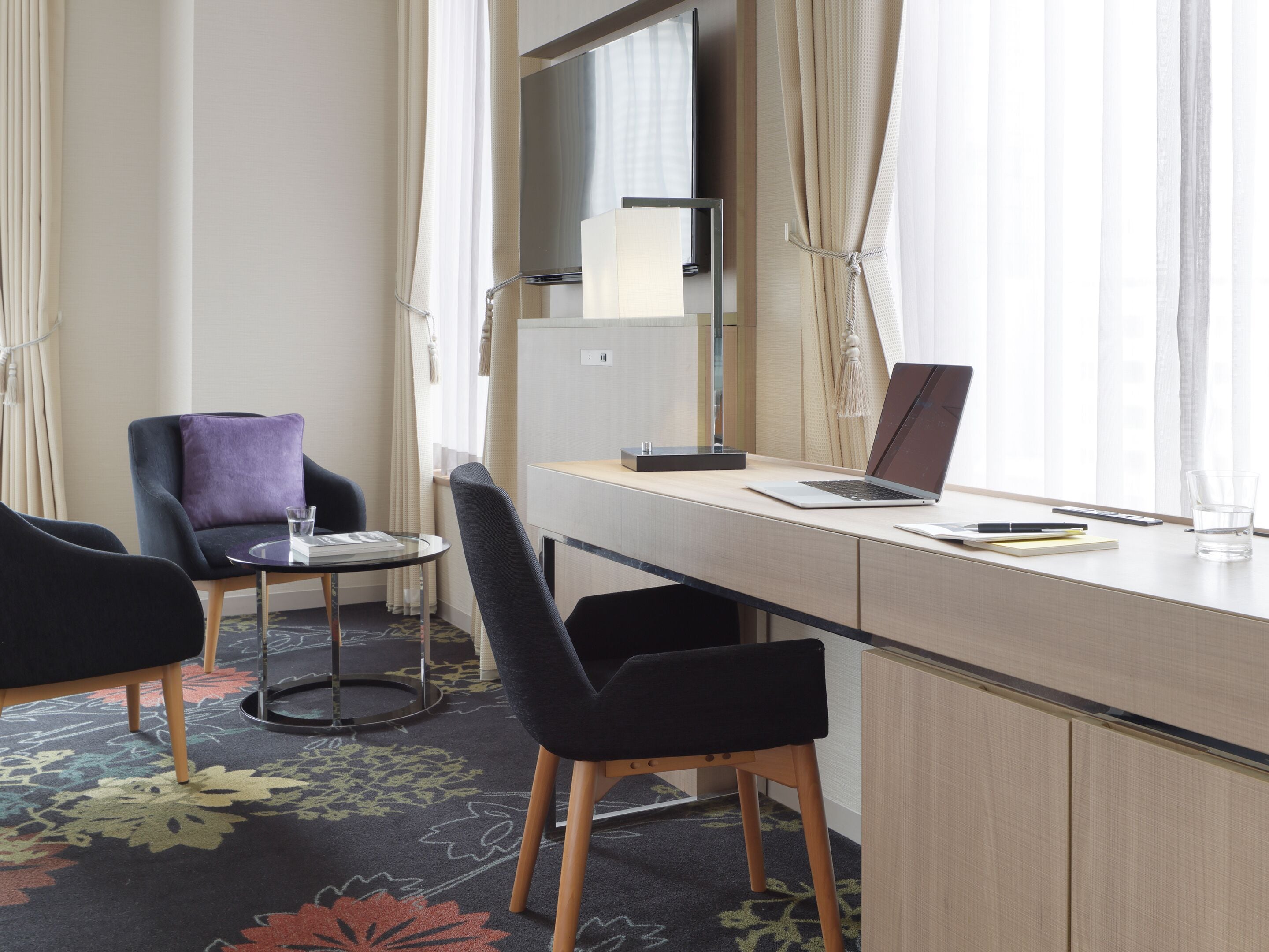 A hotel room with a desk, two chairs, a laptop, a glass of water, and a small table with a glass.