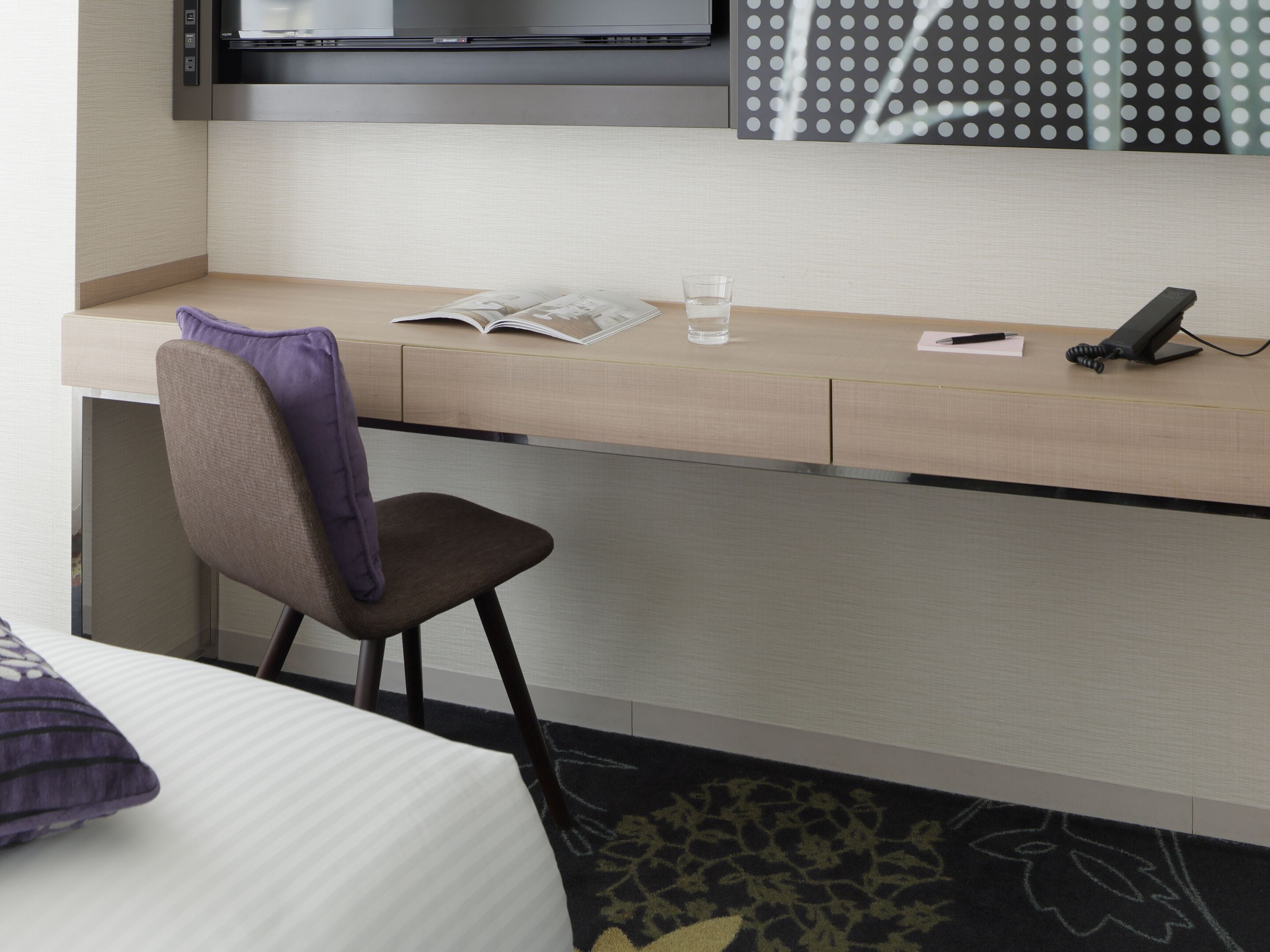 A hotel room with a bed, a desk with a chair, a television, and a patterned wall.