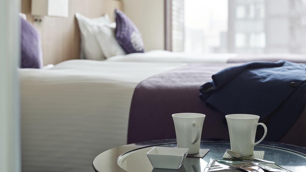A hotel room with a bed, two white cups, a small tray, and a jacket on a chair.