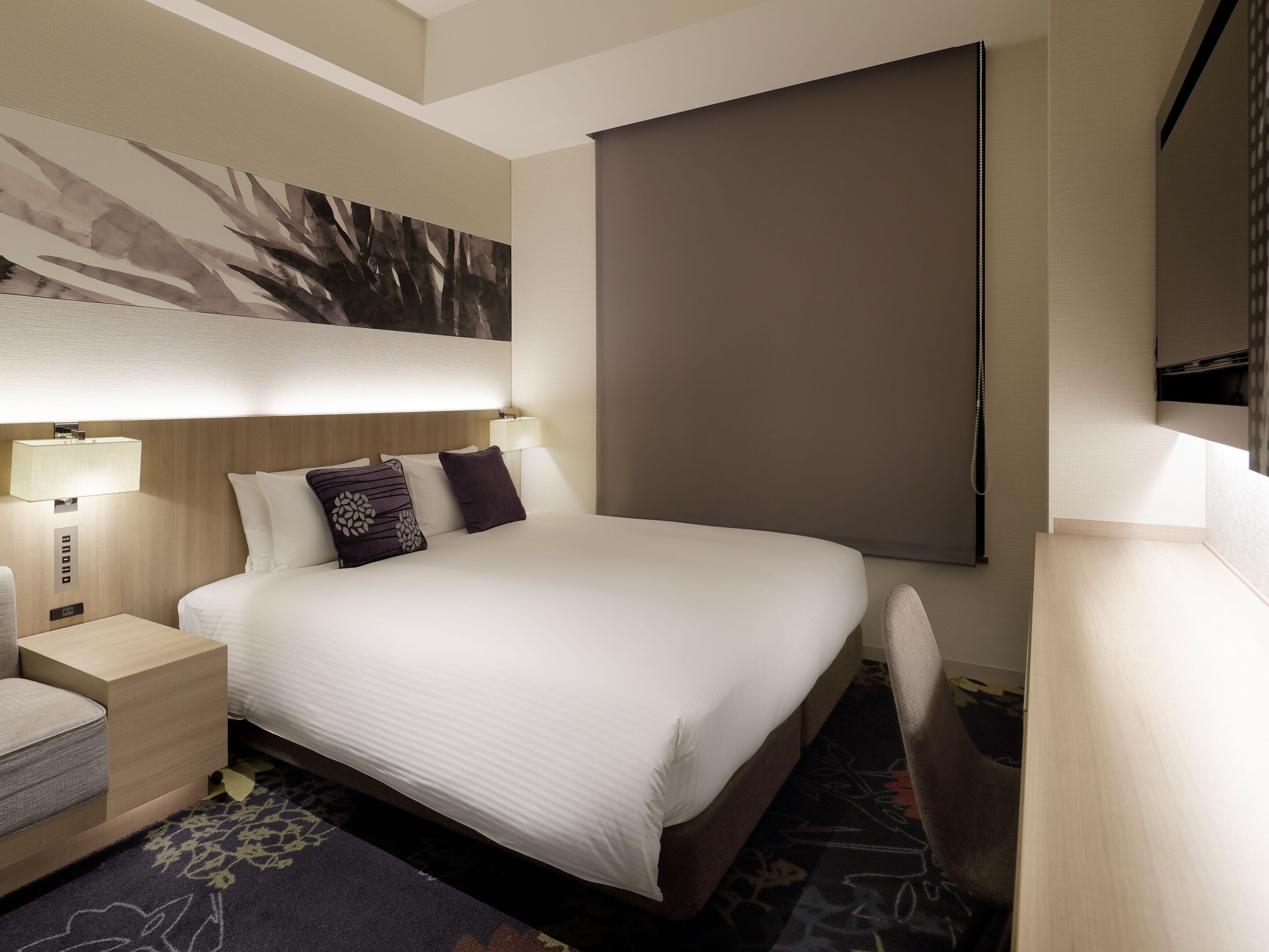 A hotel room with a large bed, bedside tables, a desk, a chair, a painting on the wall, and a large window.