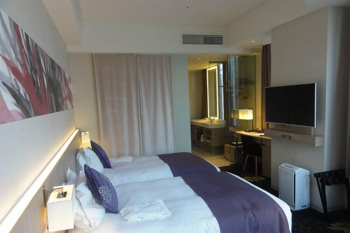 Habitación de hotel con dos camas, un escritorio, una silla, un televisor y un baño visible a través de una puerta de cristal.