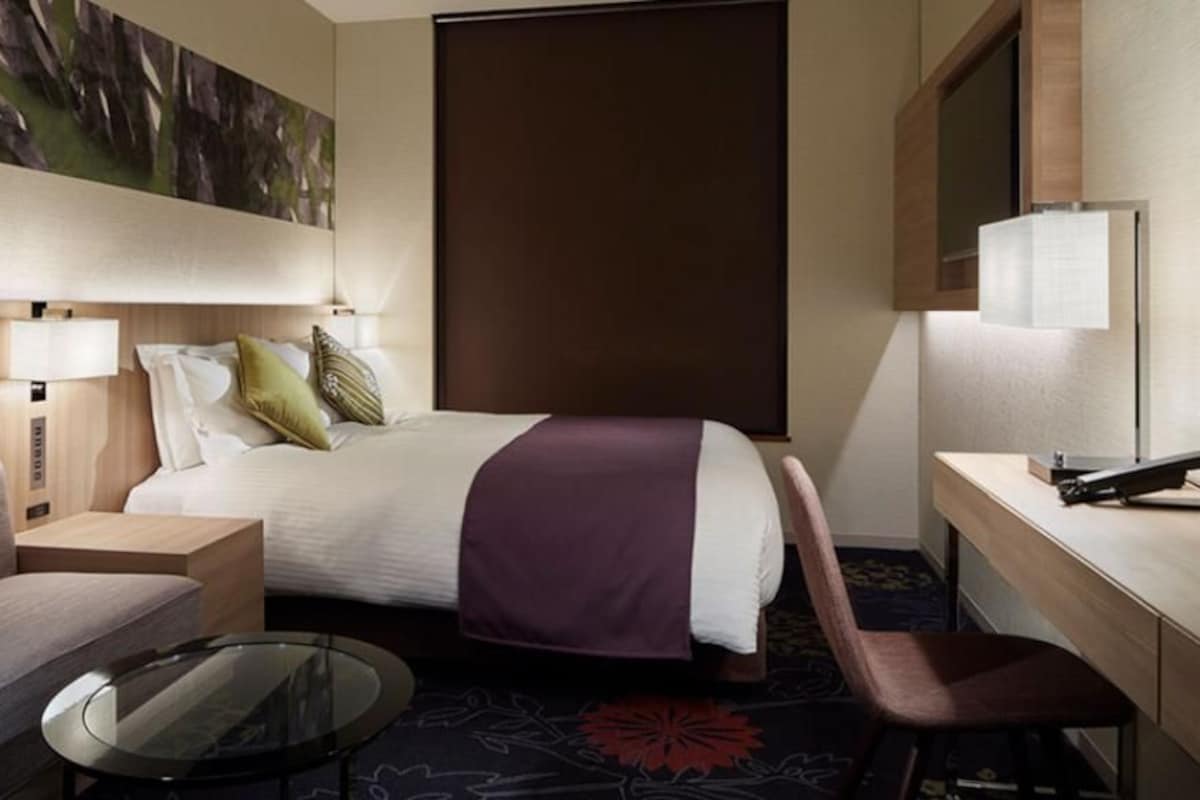 A hotel room with a bed, a desk, a chair, a sofa, a small table, and a wall-mounted TV.