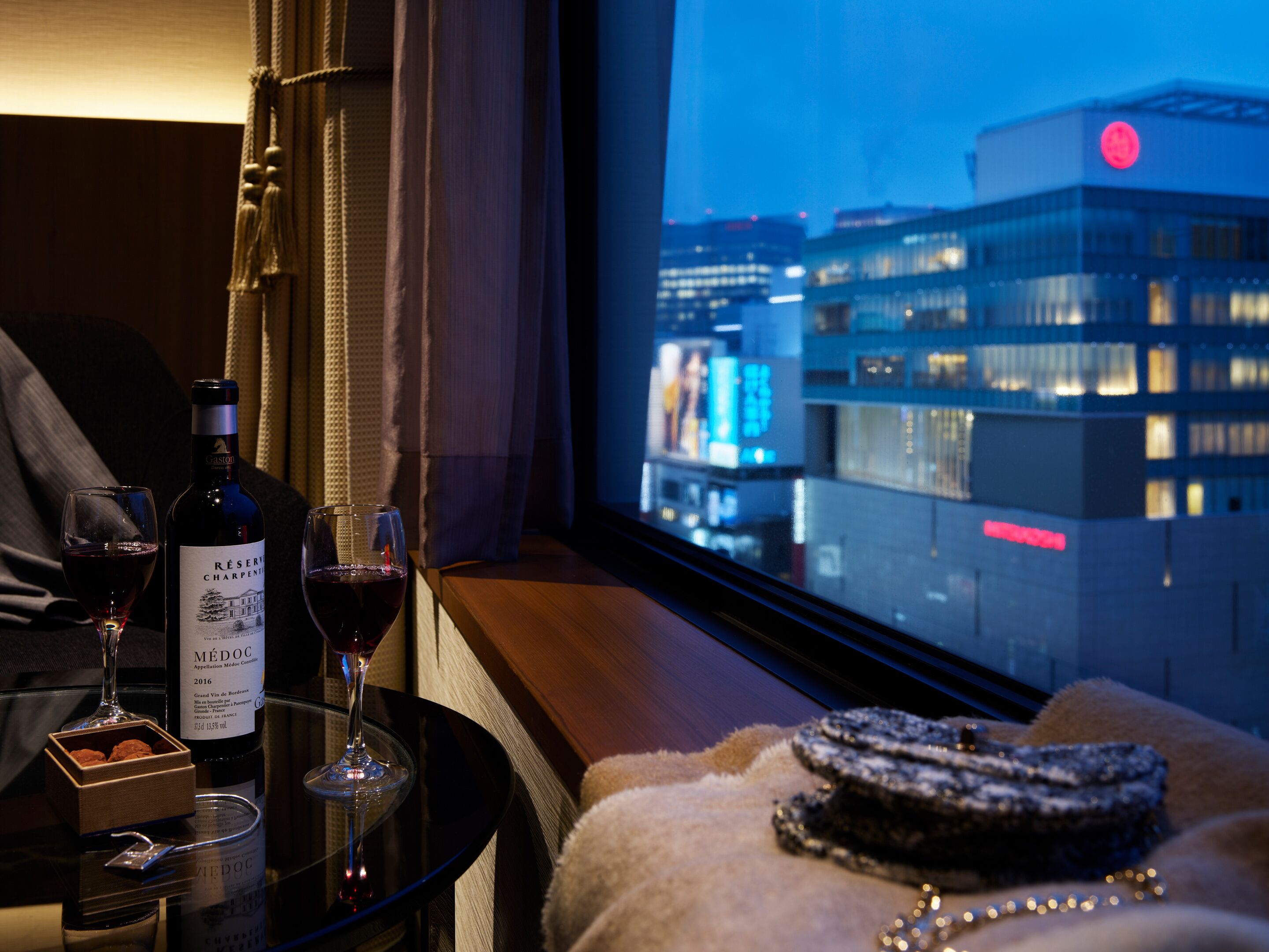 A hotel room with a view of a cityscape at night, a bottle of wine, two wine glasses, and a dessert on a table.