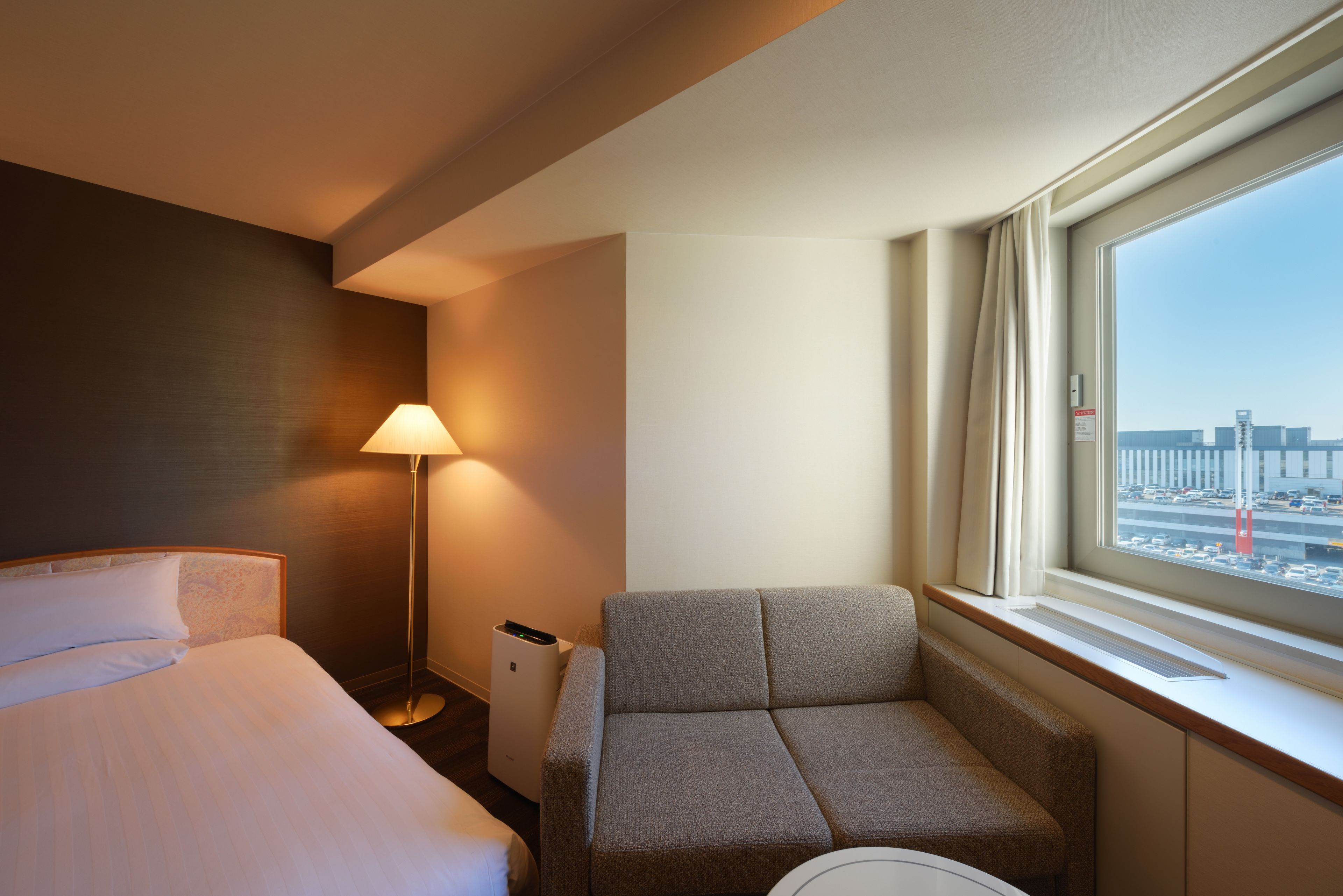 A hotel room with a bed, a sofa, a lamp, and a window with a view of a cityscape.