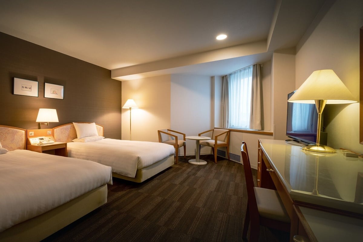 Une chambre d’hôtel avec deux lits, un bureau, une chaise, une télévision et une fenêtre avec des rideaux.