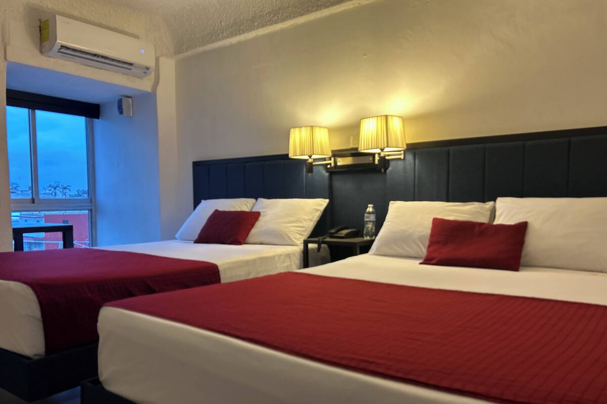 Habitación de hotel con dos camas, cada una con ropa de cama roja y almohadas blancas, una mesita de noche con lámpara y un equipo de aire acondicionado en la pared.