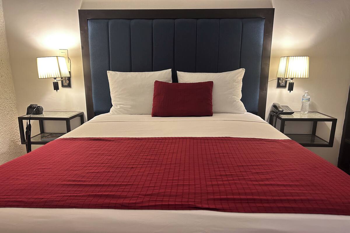 Habitación de hotel con una cama grande, una colcha acolchada roja, dos mesitas de noche con lámparas y un cabecero azul oscuro.