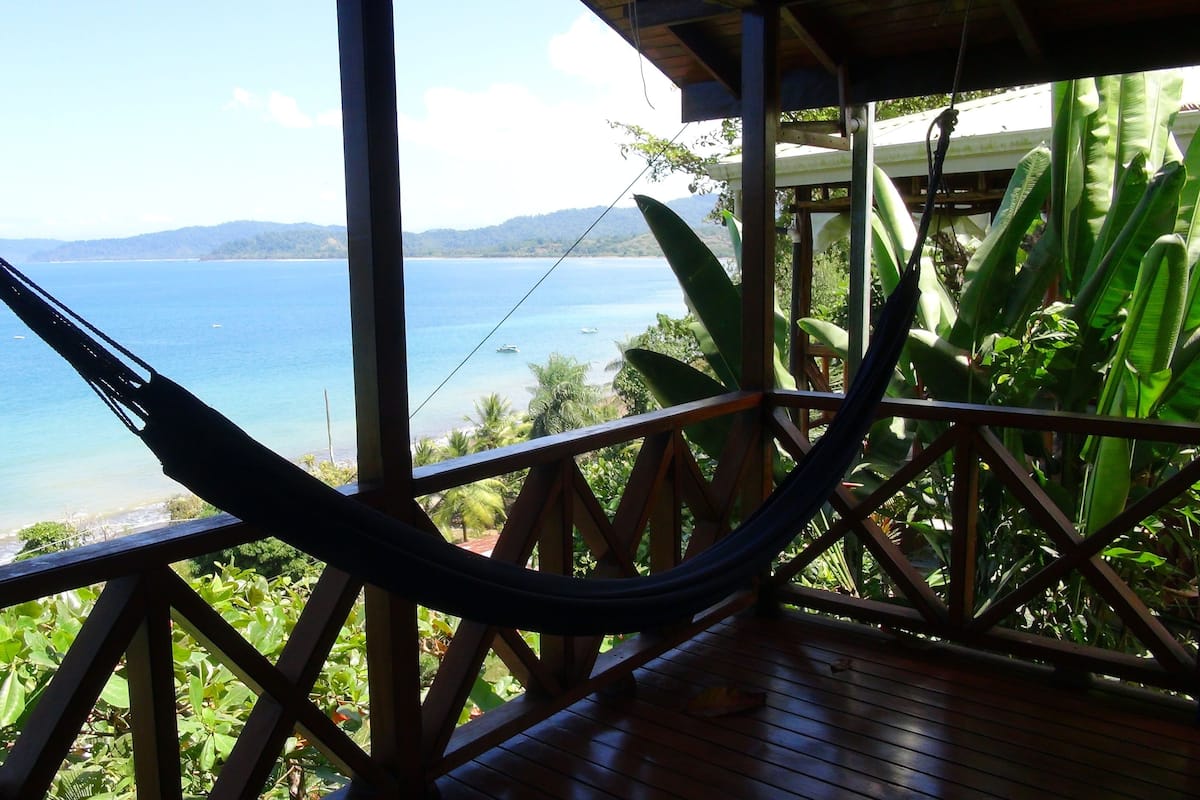 A hammock on a wooden deck overlooking a beach with boats and palm trees.