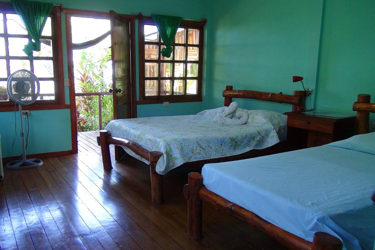 A room with two wooden beds, a wooden desk, a small table, a fan, and a refrigerator.