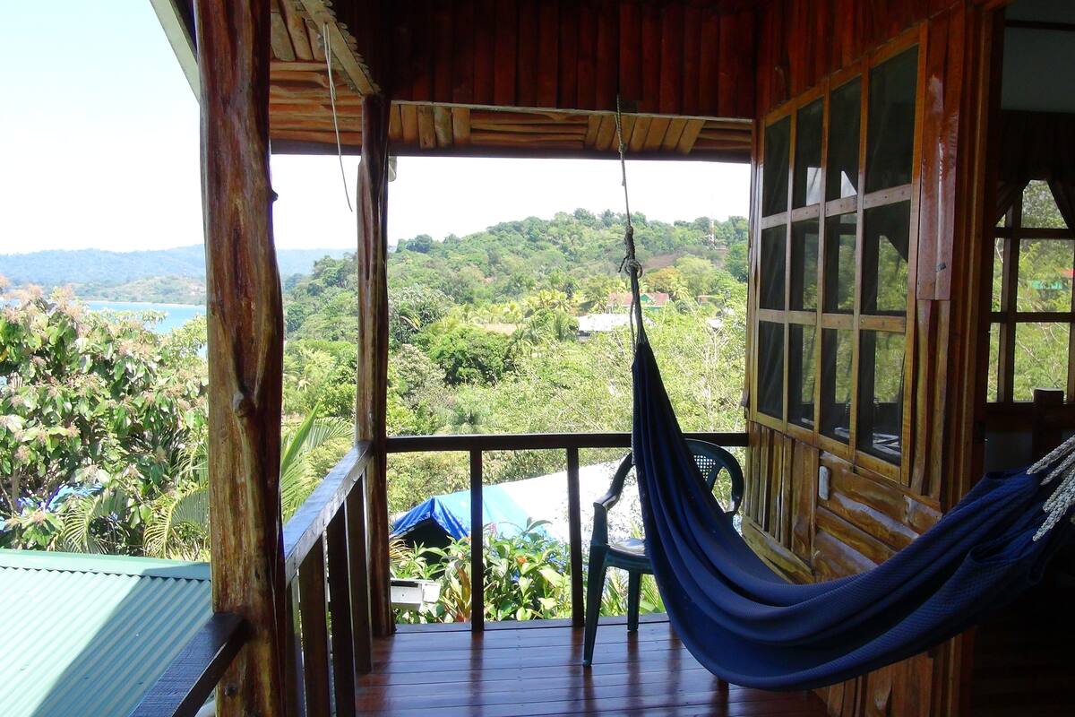 A wooden balcony with a hammock, a view of a lush landscape, and a clear sky.