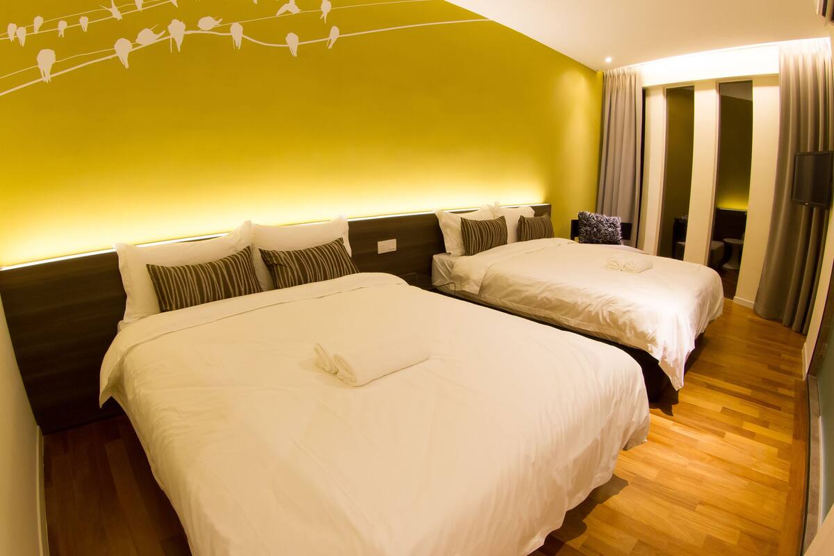 A hotel room with two beds, a wooden floor, a green accent wall, and a sliding door leading to a bathroom.