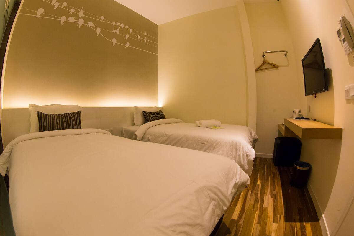 A hotel room with two beds, a wooden floor, a wall-mounted TV, and a small desk with a chair.