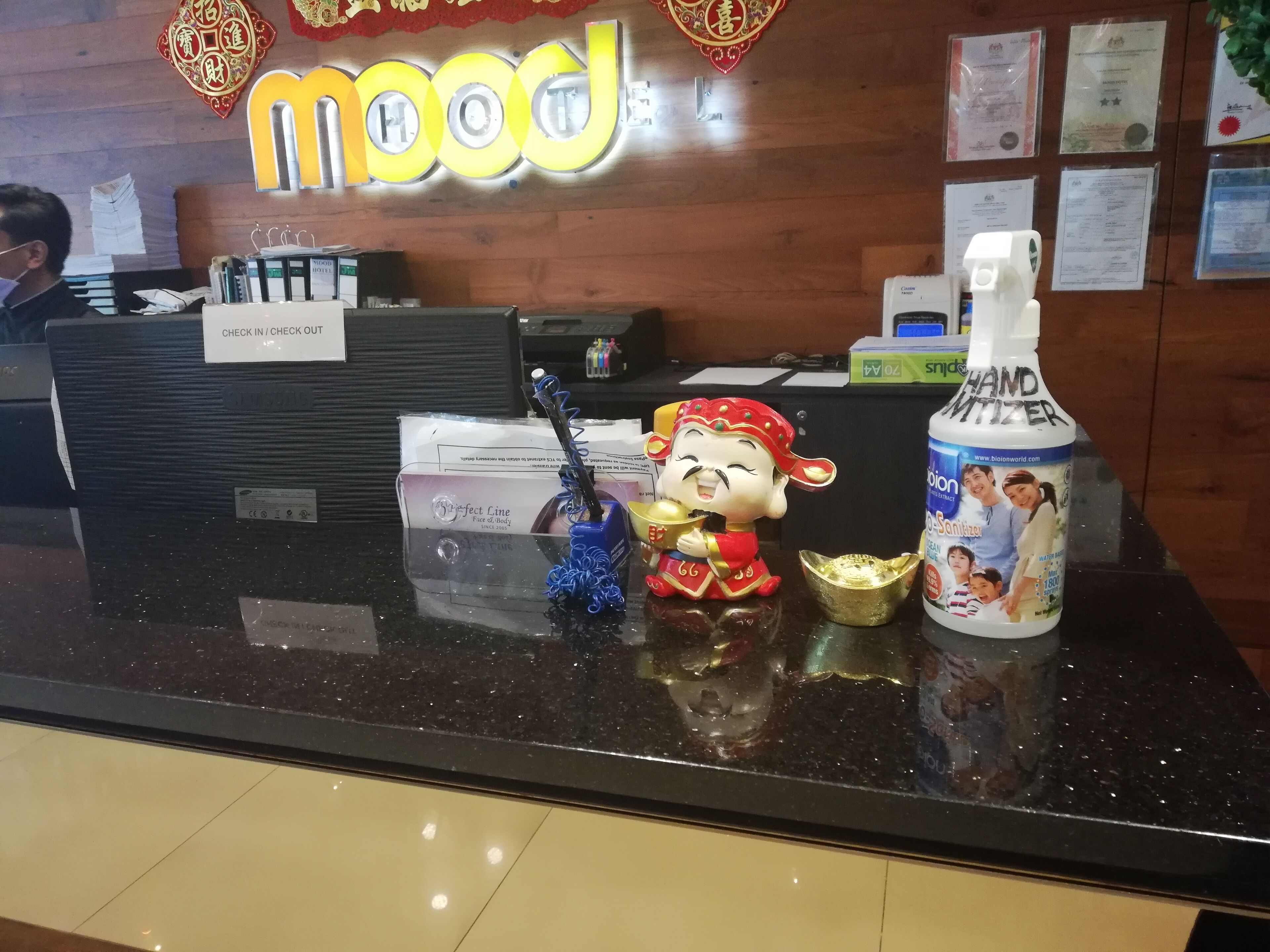 A reception desk with a 'Mood' sign, a hand sanitizer dispenser, a 'Perfect Line' face and body wash, and a decorative figurine.