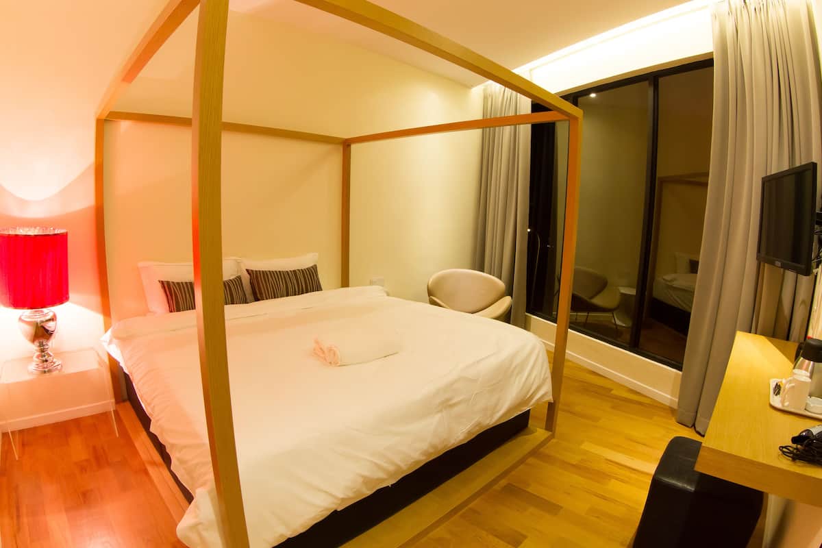 A four-poster bed with white linens, a bedside table with a red lamp, a chair, and a desk with a coffee maker.