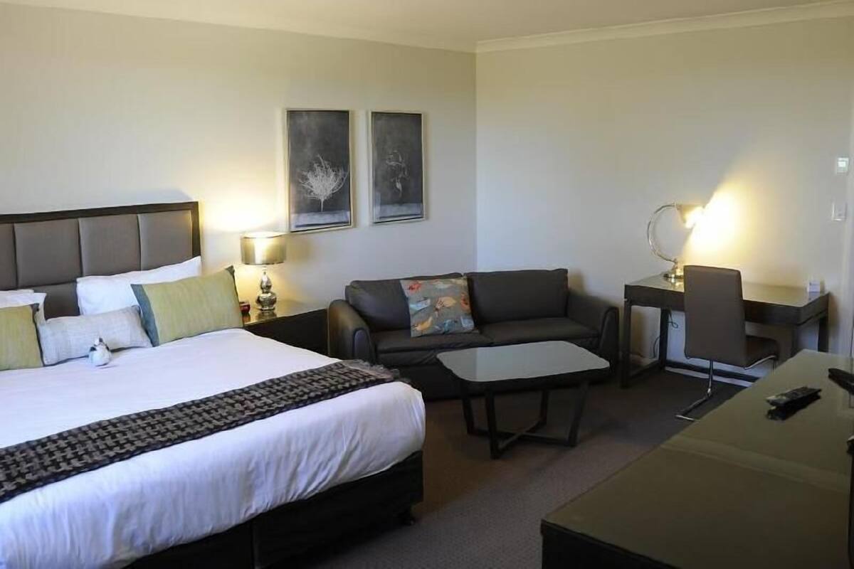 Habitación de hotel con cama, sofá, escritorio y silla. Dos lámparas de pared e iluminación en el techo.