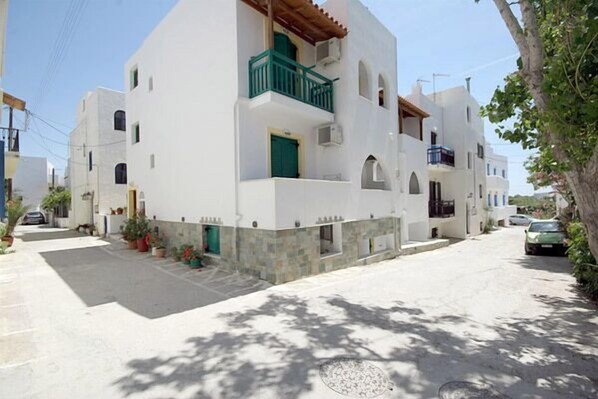 A white building with a green door and balcony, surrounded by a narrow street and parked cars. - Studios Simos (Naxos)