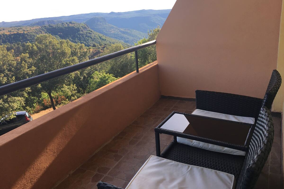 A balcony with a wicker chair and a small table, overlooking a mountainous landscape.