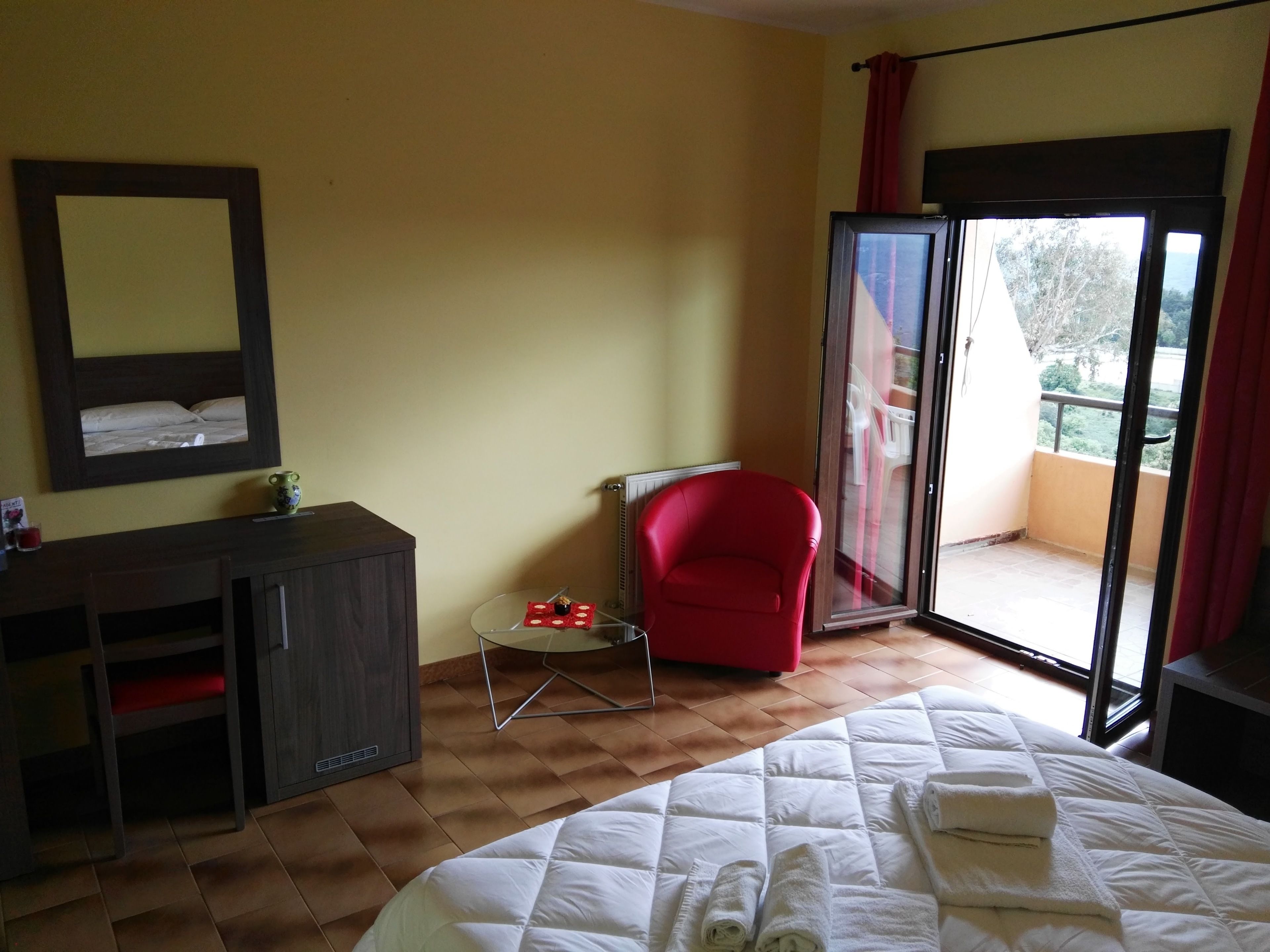 Habitación de hotel con cama, una silla roja, una mesita, un espejo y una puerta corrediza de vidrio que da a un balcón.