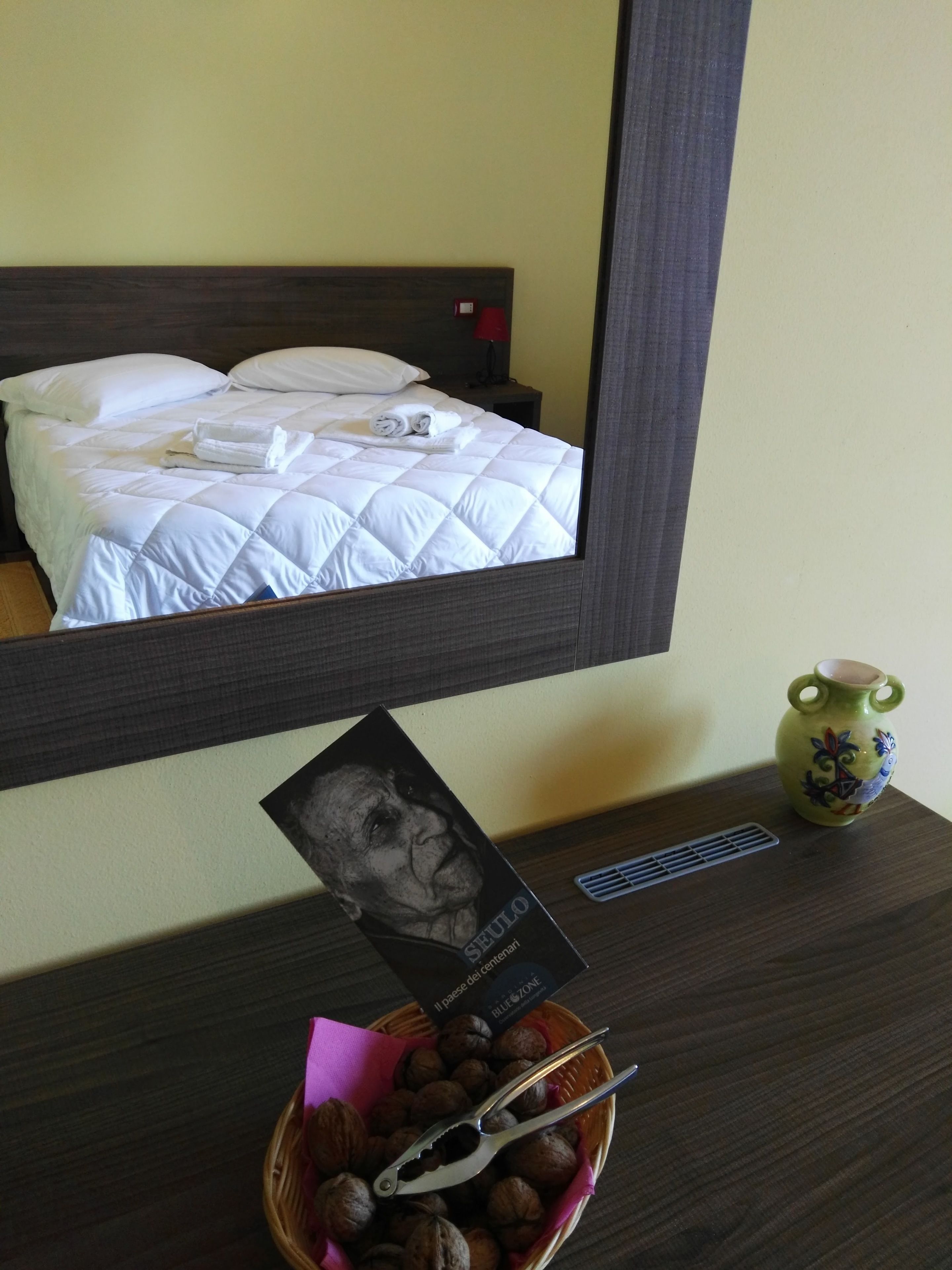 A neatly made bed with a white duvet and pillows, a small vase with a floral design, and a basket of nuts with a pair of tongs on a wooden nightstand.