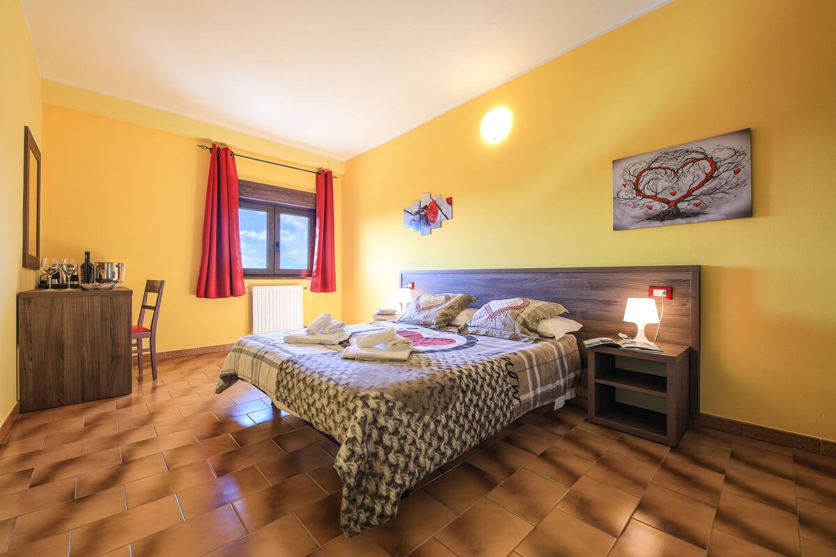 A bedroom with a bed, bedside tables, a nightstand, a window with curtains, a wall clock, and a painting.