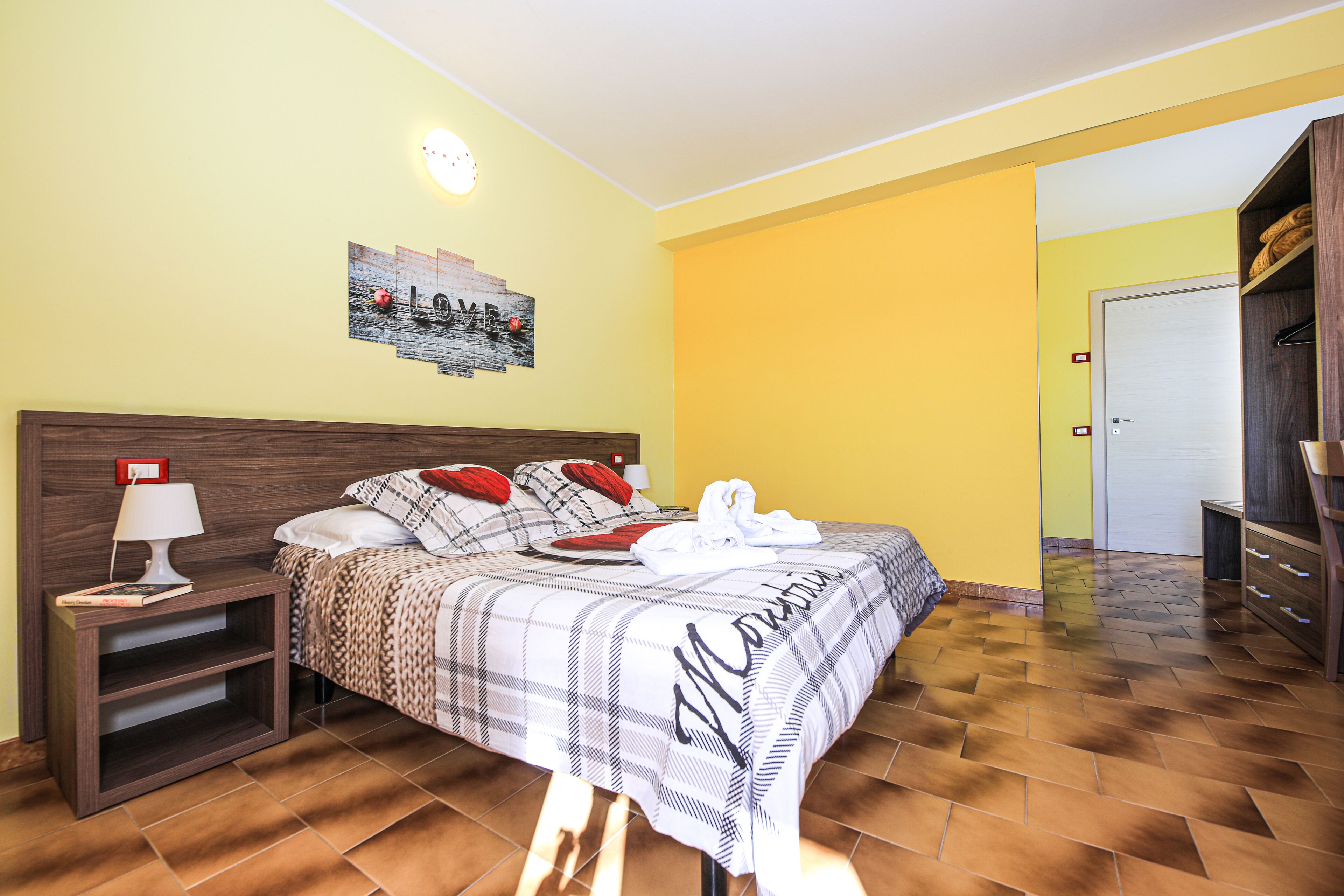 Quarto de hotel com cama, mesa de cabeceira, abajur, um quadro na parede e guarda-roupa de madeira.