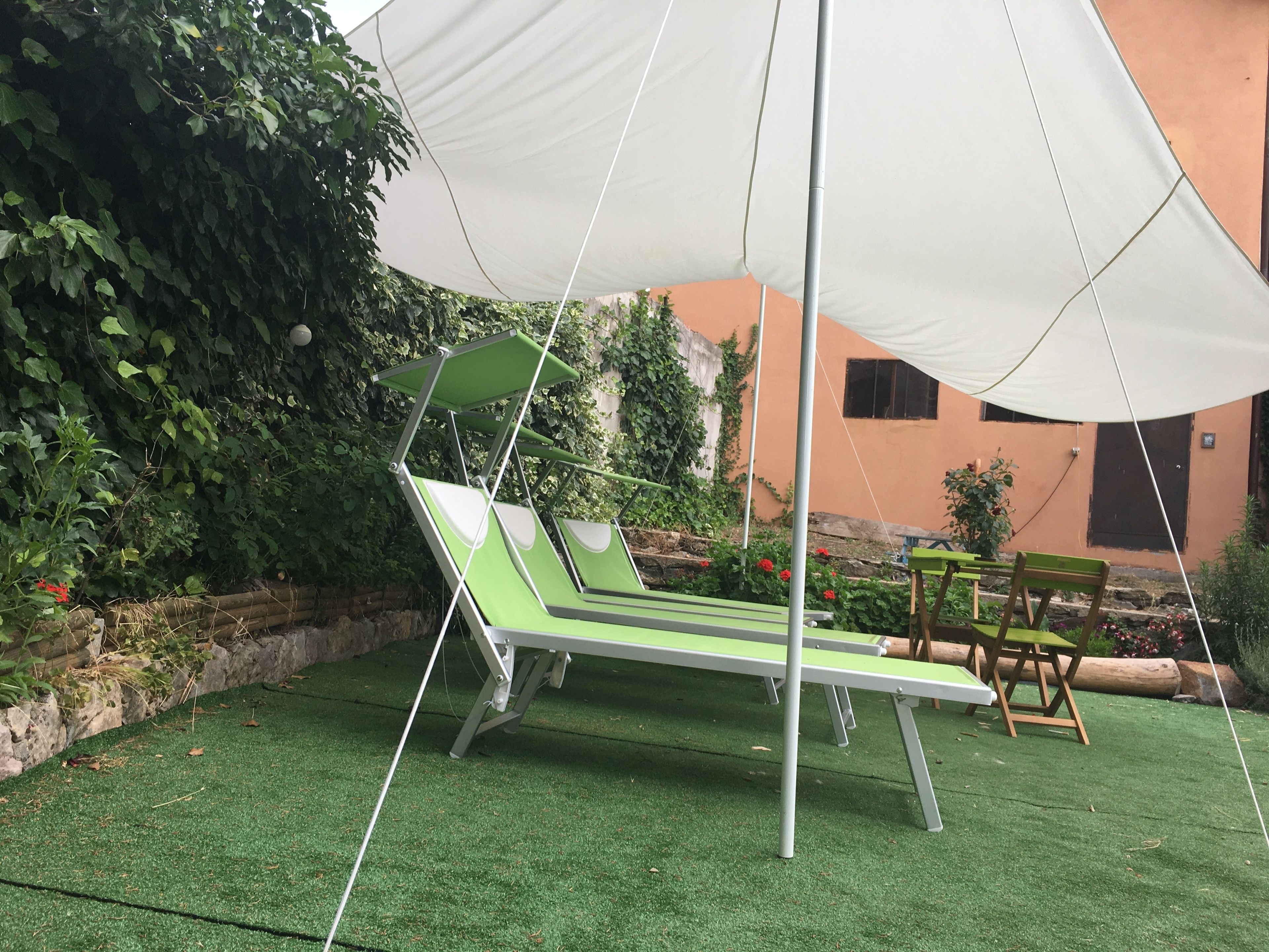 Un patio con dos sillones verdes bajo un toldo blanco, rodeado de vegetación y un edificio con pared naranja.