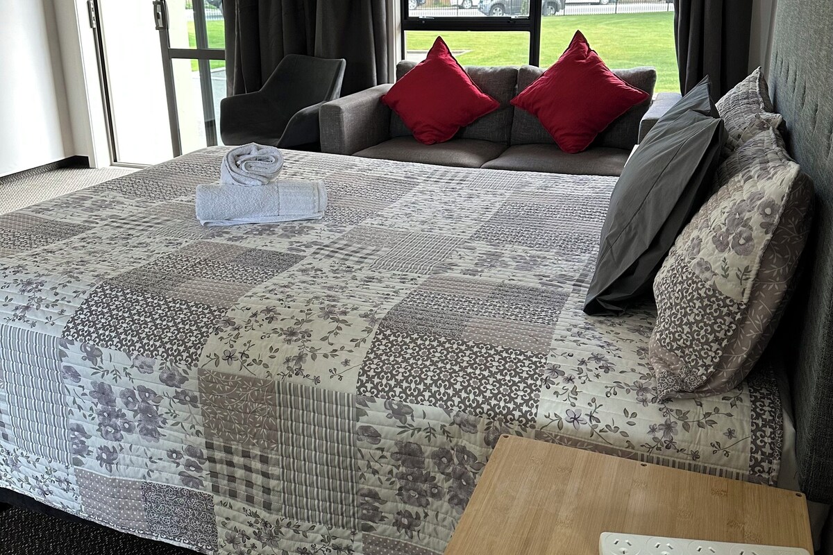 A bedroom with a large bed, a wooden nightstand, a grey sofa with red cushions, and a window with a view of the outside.