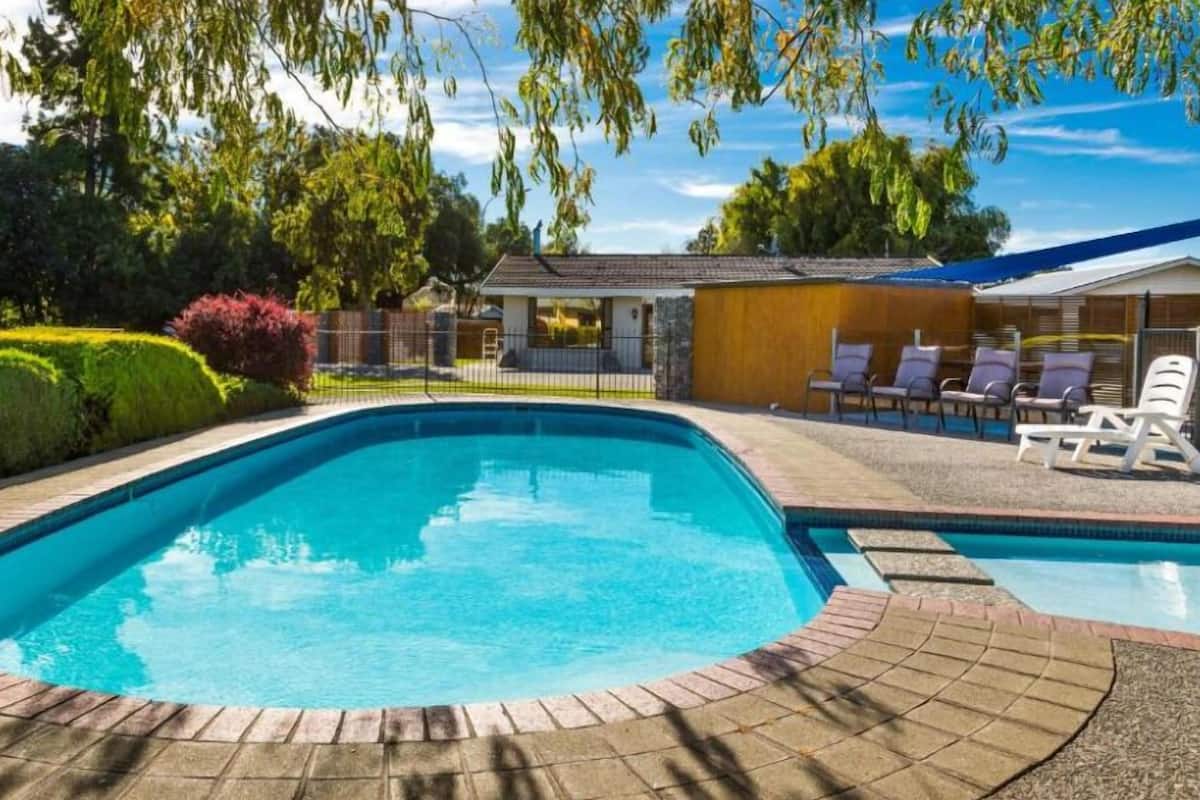 A rectangular swimming pool with a brick border, surrounded by a patio area with lounge chairs and a covered seating area.