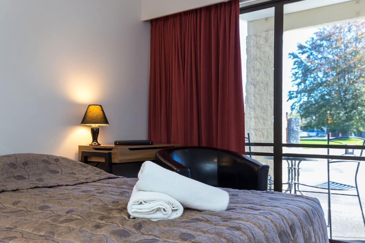 A hotel room with a bed, a desk, a chair, a lamp, and a view of the outdoors.