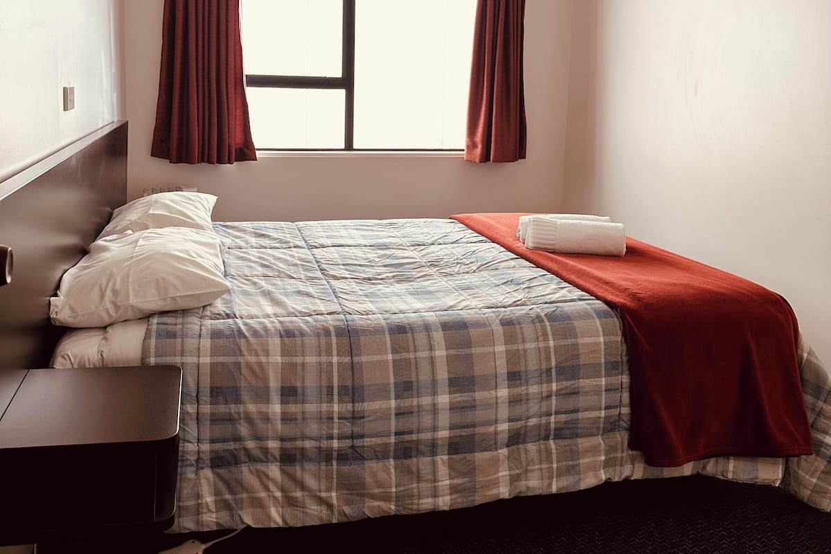 A neatly made bed with a plaid bedspread and a red blanket, a bedside table, and a power outlet.