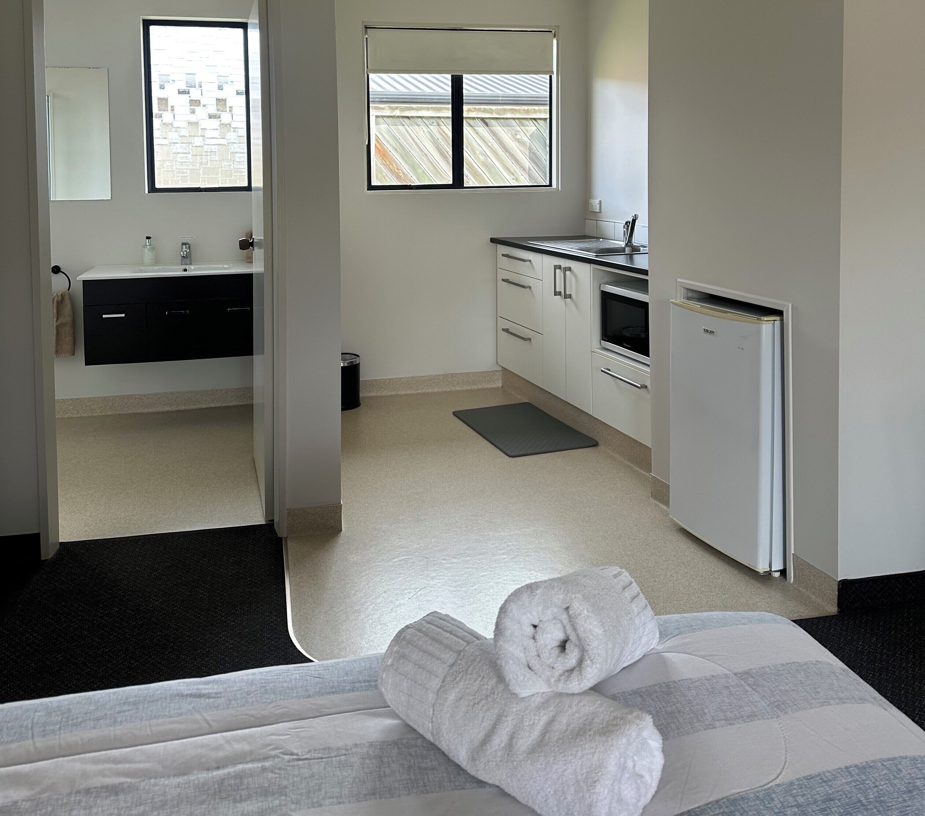 Une chambre d’hôtel comprenant un lit, une salle de bain avec un lavabo et un miroir, une cuisine équipée d’un réfrigérateur et d’une cuisinière, et une fenêtre donnant sur l’extérieur.