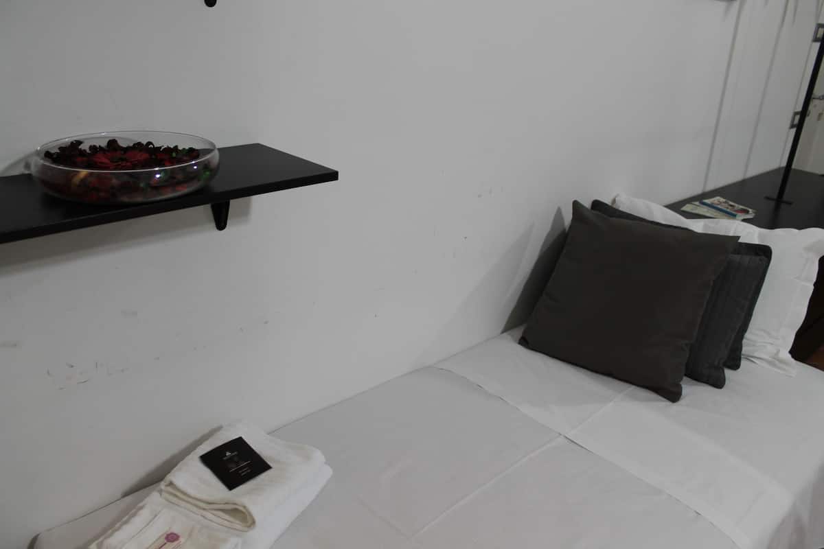 Ein ordentlich bezogenes Bett mit weißen Laken und dunklen Kissen, ein kleiner, schwarzer Nachttisch mit einer Zeitschrift und eine Schale mit roten Beeren auf einem schwarzen Regal über dem Bett.
