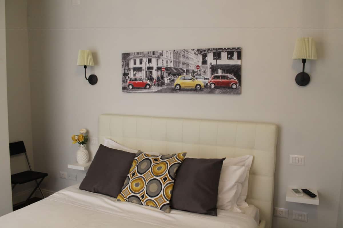 Ein Hotelzimmer mit Bett, Nachttisch, Lampe, einer Vase mit Blumen und Wandbildern von Autos auf einer Straße.