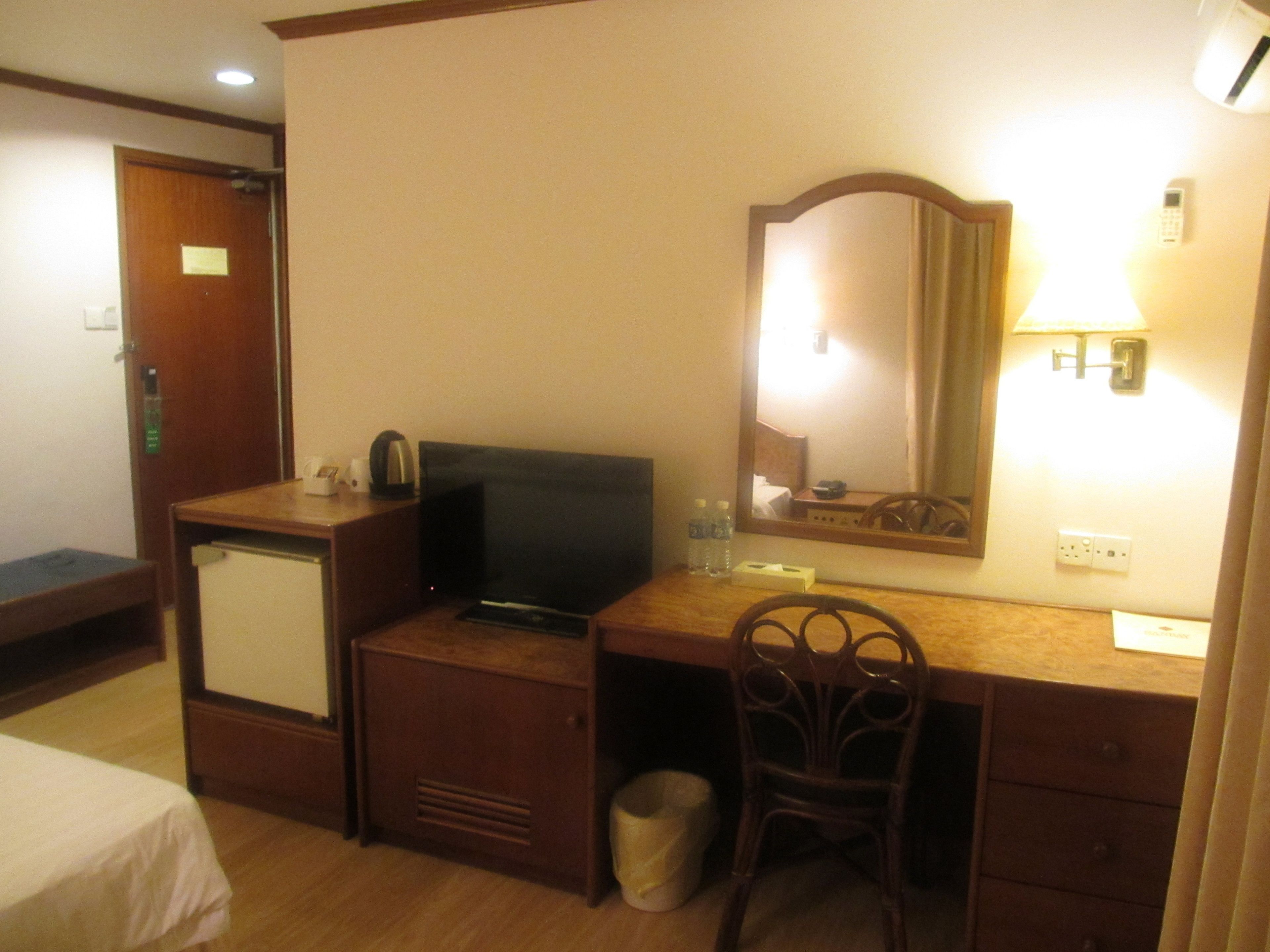 A hotel room with a wooden desk, a television, a chair, a small trash bin, and a bed.