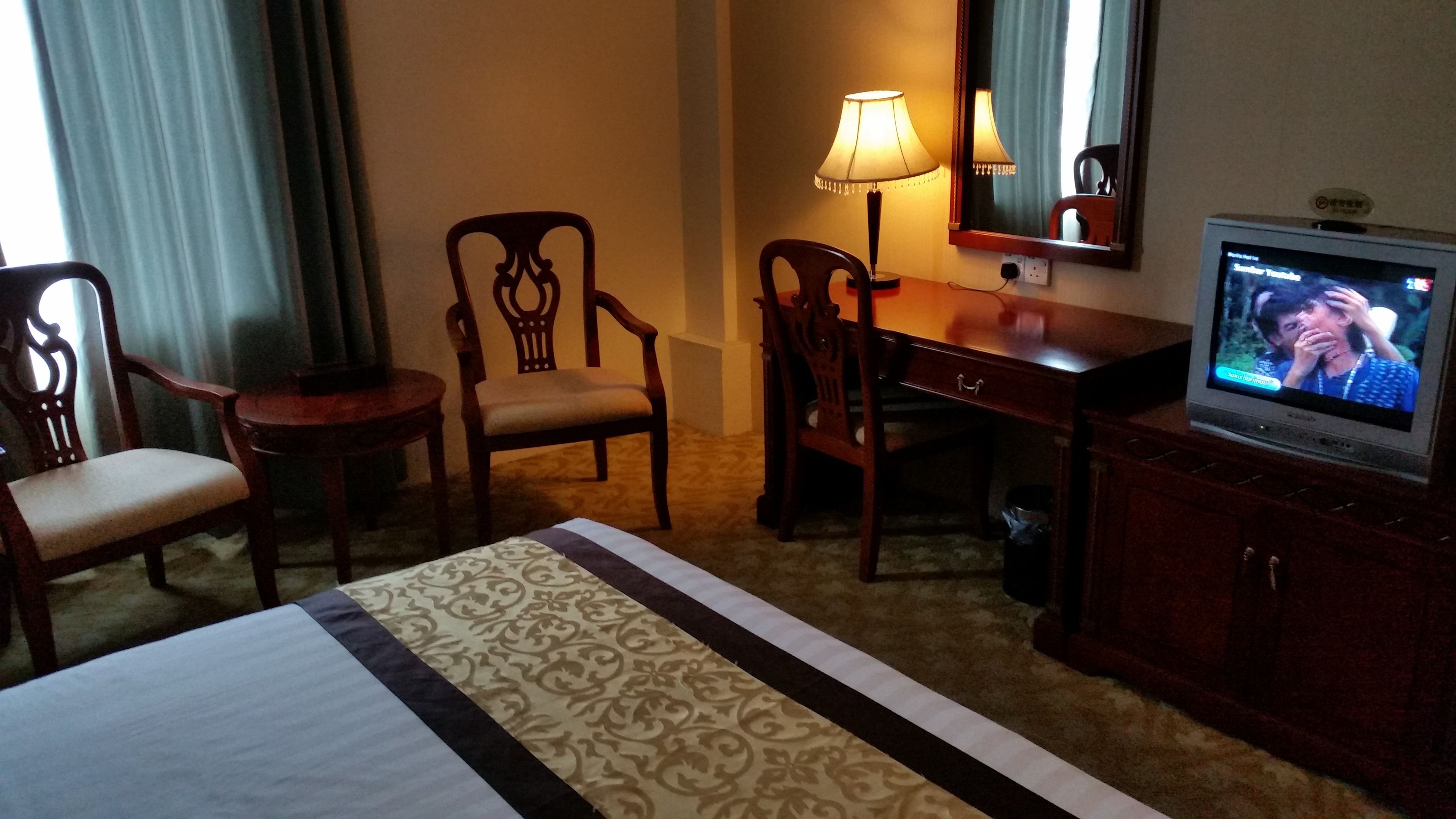 A hotel room with a bed, two wooden chairs, a desk with a lamp, a television, and a mirror.