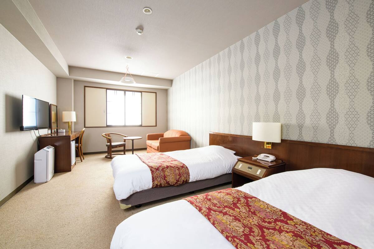 A hotel room with two beds, a desk, a chair, a TV, and a window.