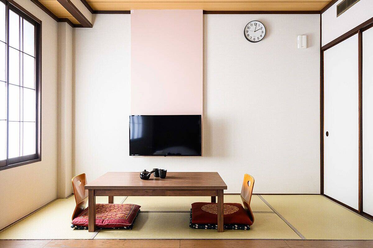 A traditional Japanese room with tatami flooring, a low table with cushions, a television, and a clock on the wall.
