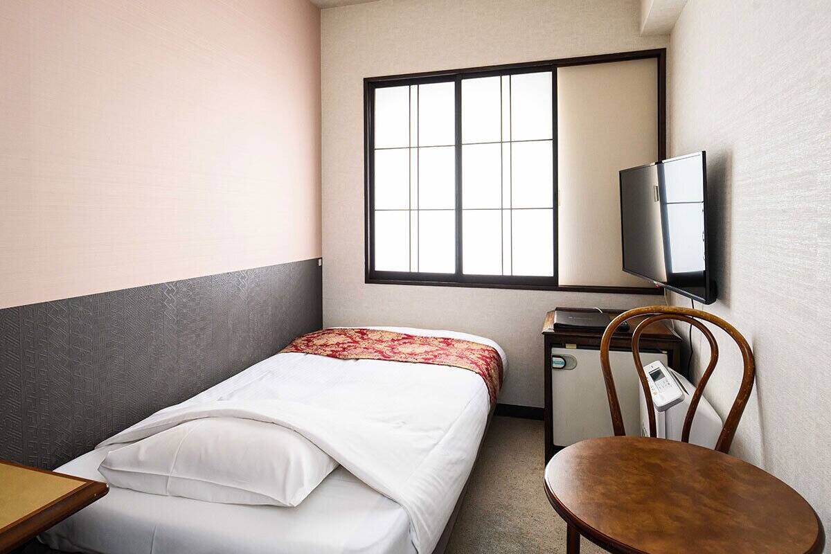 A small hotel room with a single bed, a desk, a chair, a television, and a window with blinds.