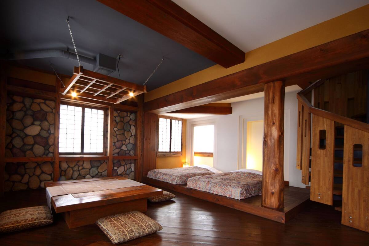 A traditional Japanese-style room with tatami flooring, a low wooden ceiling, and a stone wall. There are two tatami mats with cushions, a low wooden table, and a hanging light fixture.
