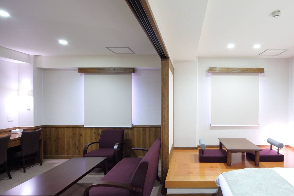 A hotel room with a wooden desk, purple chairs, a bed, and a small table.