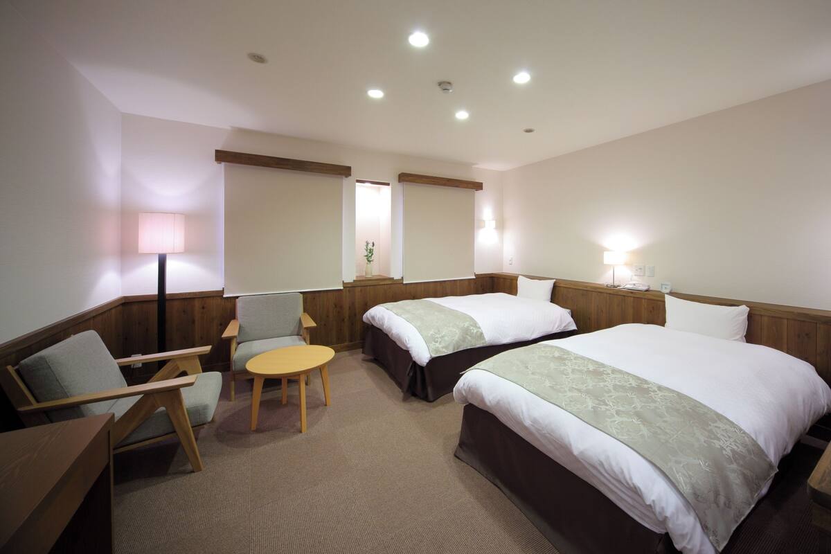 A hotel room with two beds, a chair, a small table, and a lamp.